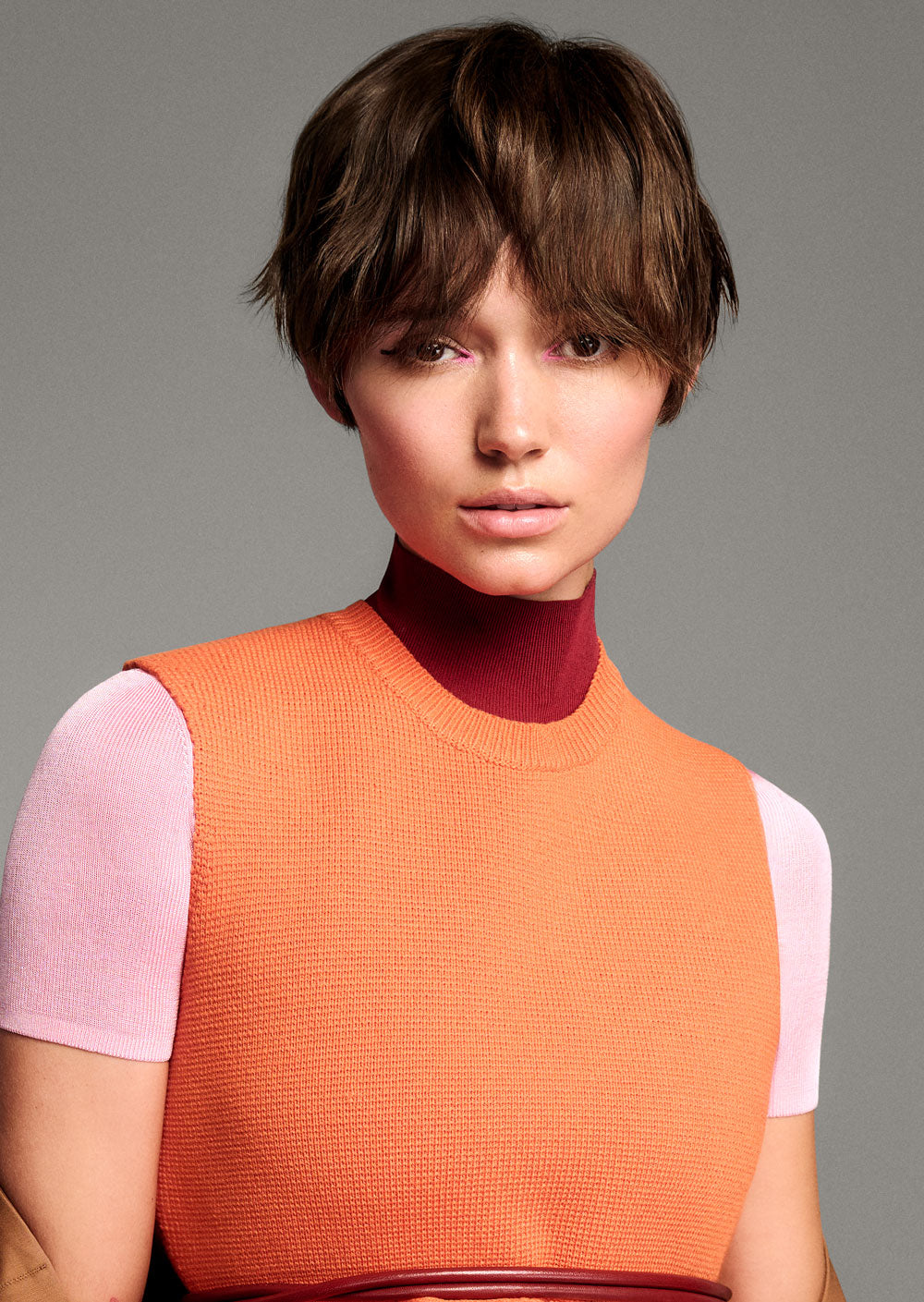 TONI & GUY EDITION ONE MIA TRIXIE CUTWoman wearing a colorful outfit with an orange vest over a pink and white top on a gray background- 