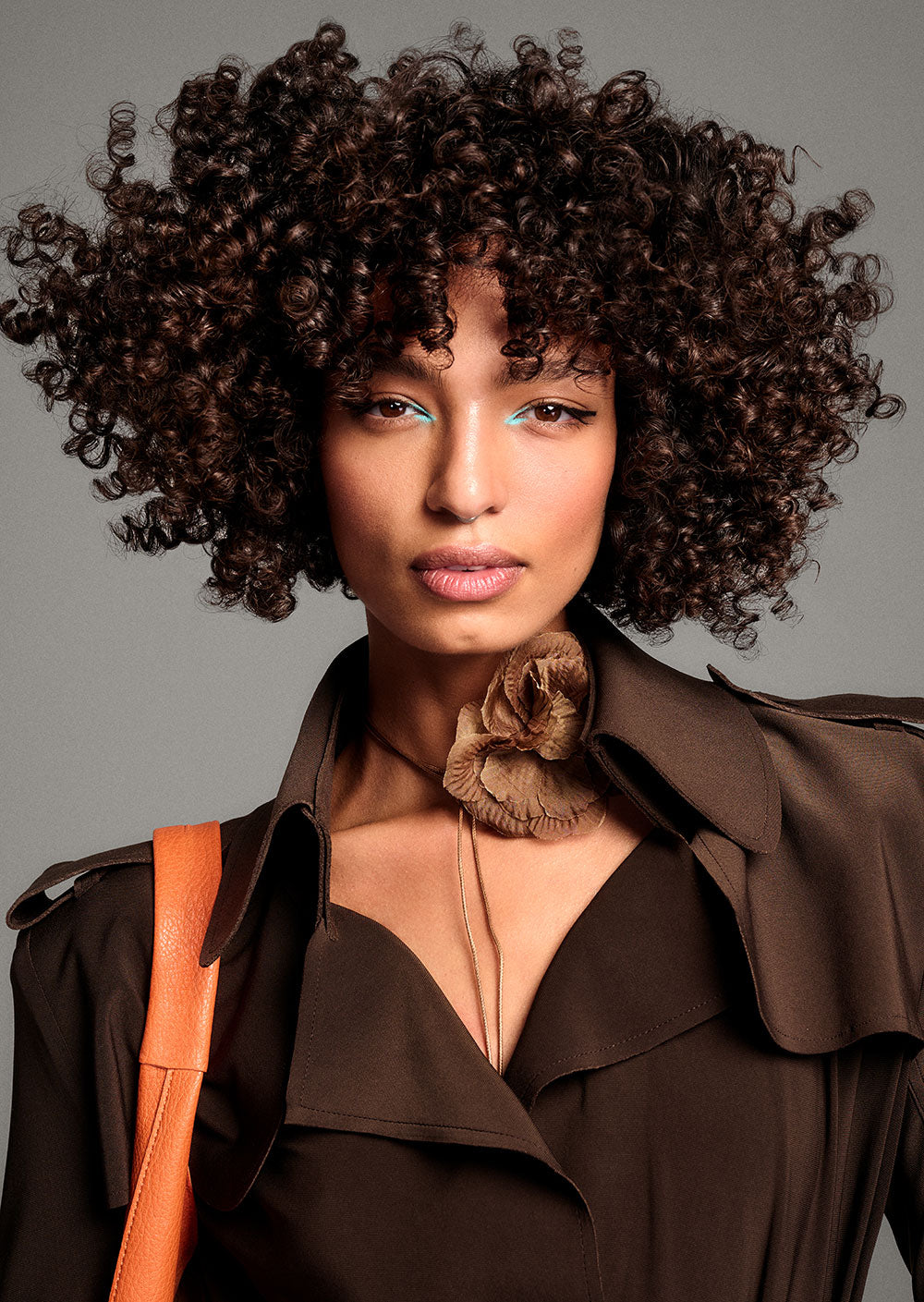 TONI & GUY EDITION ONE MARIAM HALO CUT Woman with curly hair wearing a brown coat and holding an orange bag against a gray background